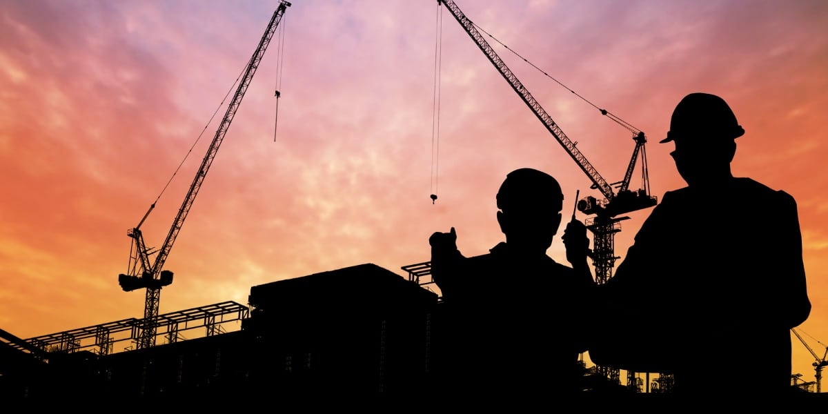 Silhouetted construction workers discussing a project at a construction site with cranes in the background, set against a vibrant sunset sky.Silhouetted construction workers discussing a project at a construction site with cranes in the background, set against a vibrant sunset sky.