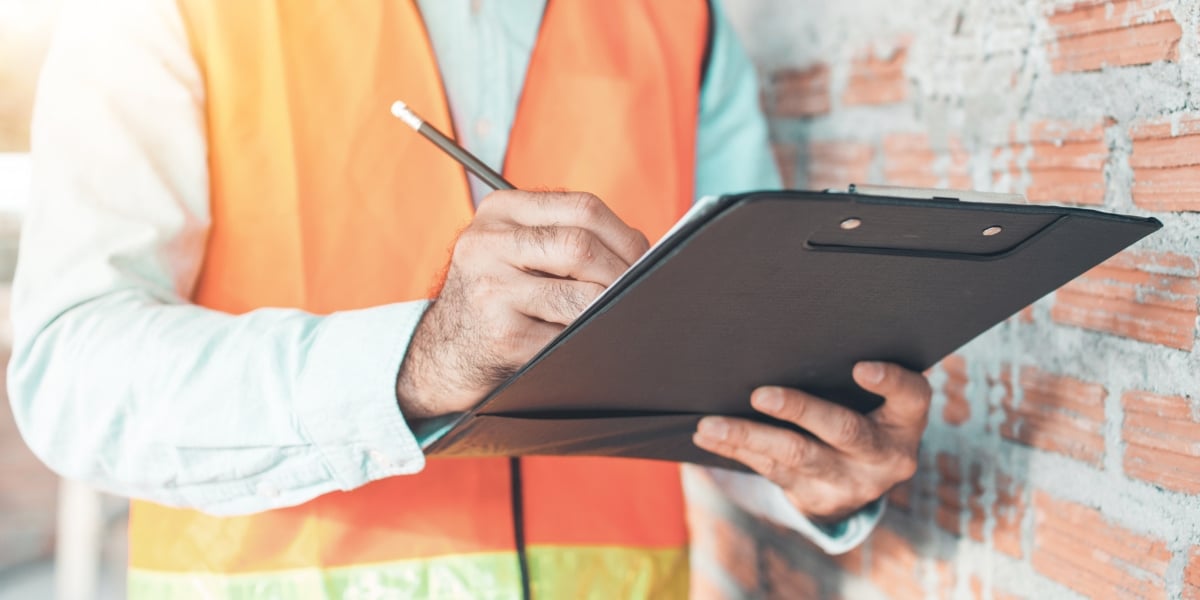 A close-up of a roofing professional wearing a high-visibility safety vest, holding a clipboard and pencil, preparing for an annual roof inspection. The background includes a partially constructed brick wall, symbolizing thorough assessment and preparation.