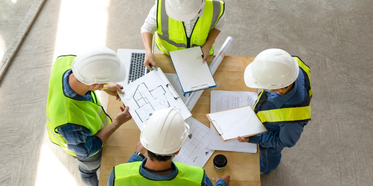 A group of commerical roofing contractors wearing high-visibility vests and helmets gathered around a table reviewing blueprints and project plans.