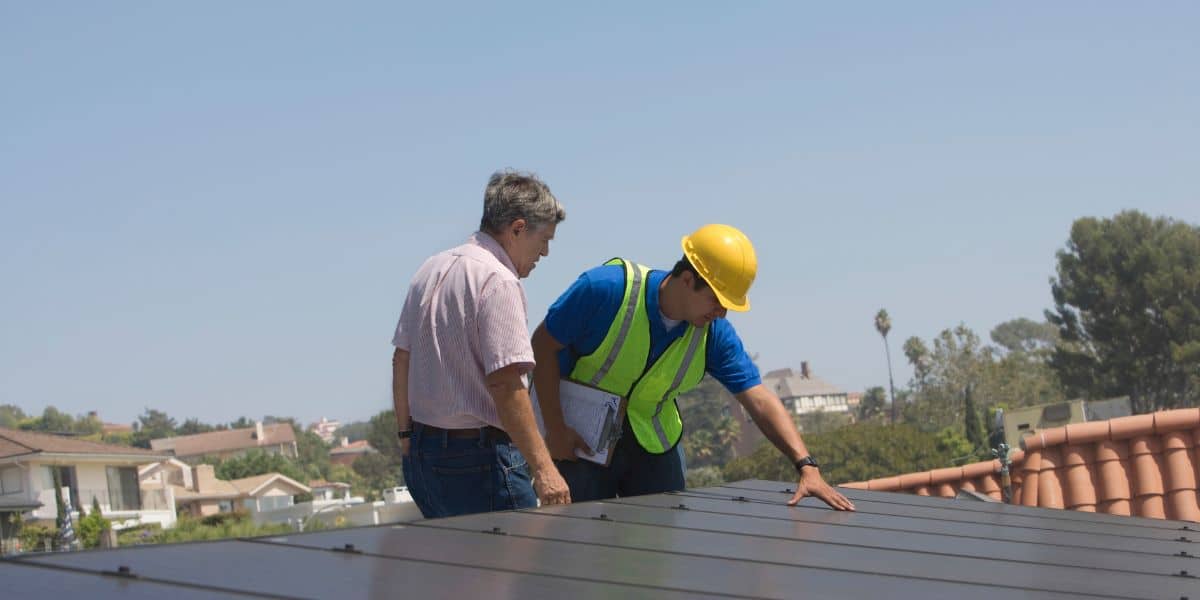 A roofing inspector wearing a hard hat and reflective vest examines a commercial roof alongside a property owner, under a clear blue sky. The scene highlights professional roof assessment for commercial properties.